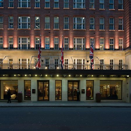 The May Fair, A Radisson Collection Hotel, Mayfair Londra Esterno foto The photo depicts a grand hotel exterior at dusk. The building has a classic architectural style, characterized by multiple stories with large windows. The facade is illuminated, highlighting the architectural features. British flags are prominently 
