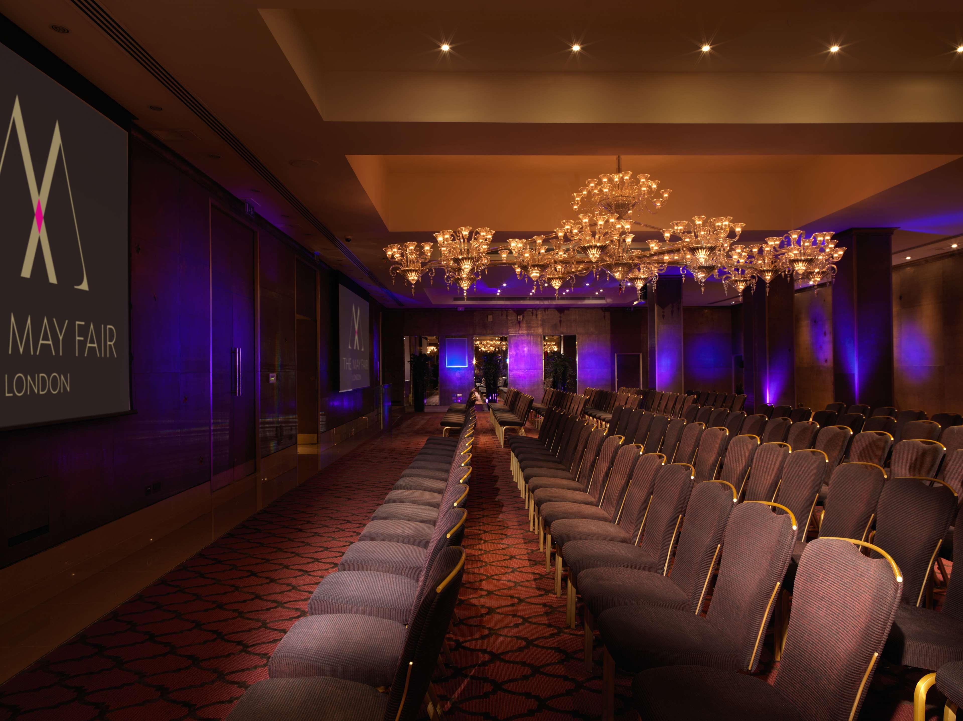 The May Fair, A Radisson Collection Hotel, Mayfair Londra Esterno foto The image shows a conference or event space with rows of elegant, upholstered chairs facing a large screen. The room is adorned with chandeliers that provide ambient lighting, and there are purple lighting accents around the space. The branding for "