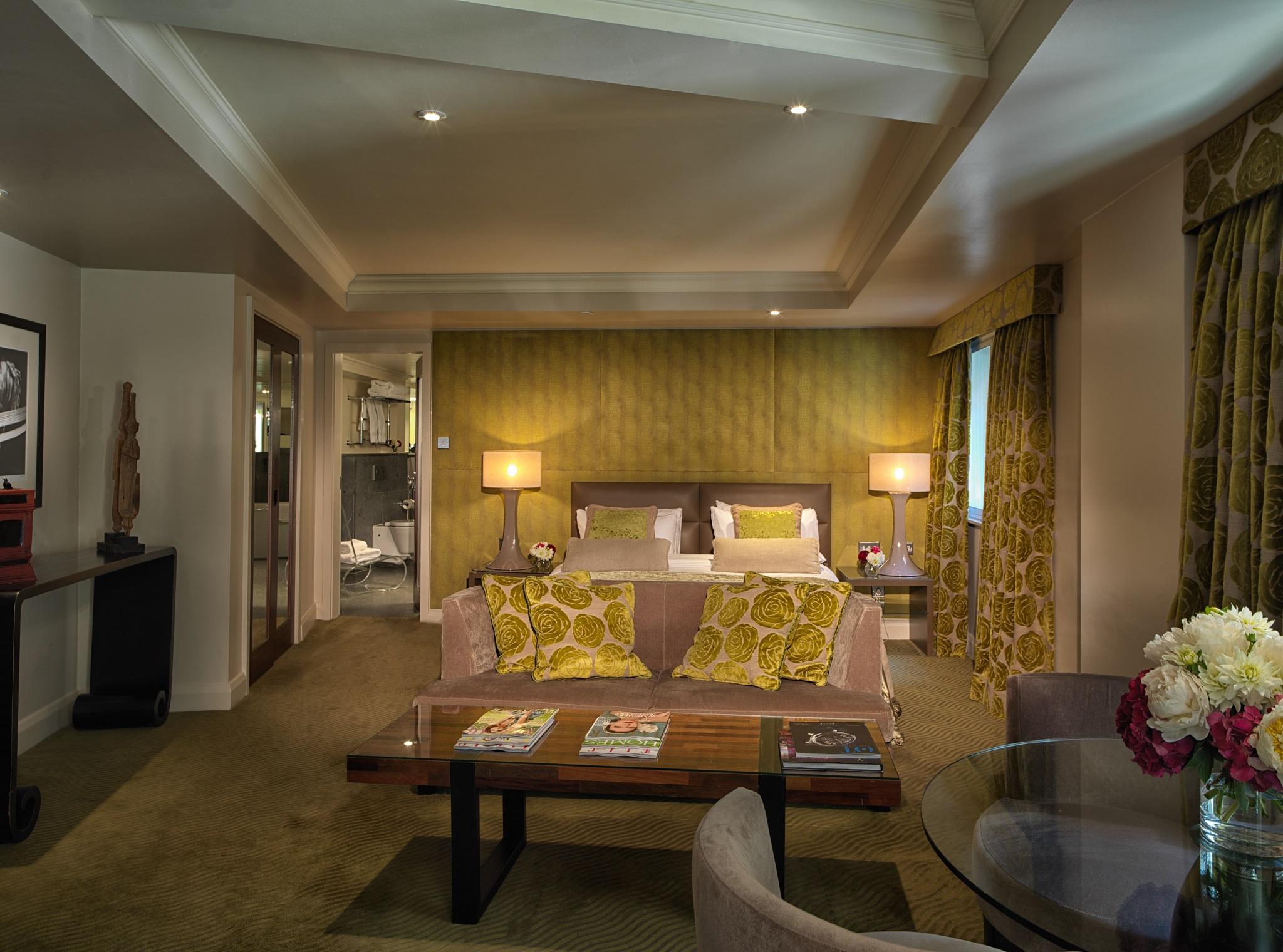 The May Fair, A Radisson Collection Hotel, Mayfair Londra Esterno foto The photo showcases a stylish hotel room or suite. It features a seating area with a couch adorned with decorative yellow pillows and a coffee table. There are two lamps on either side of the couch providing soft lighting. The walls are painted in a 