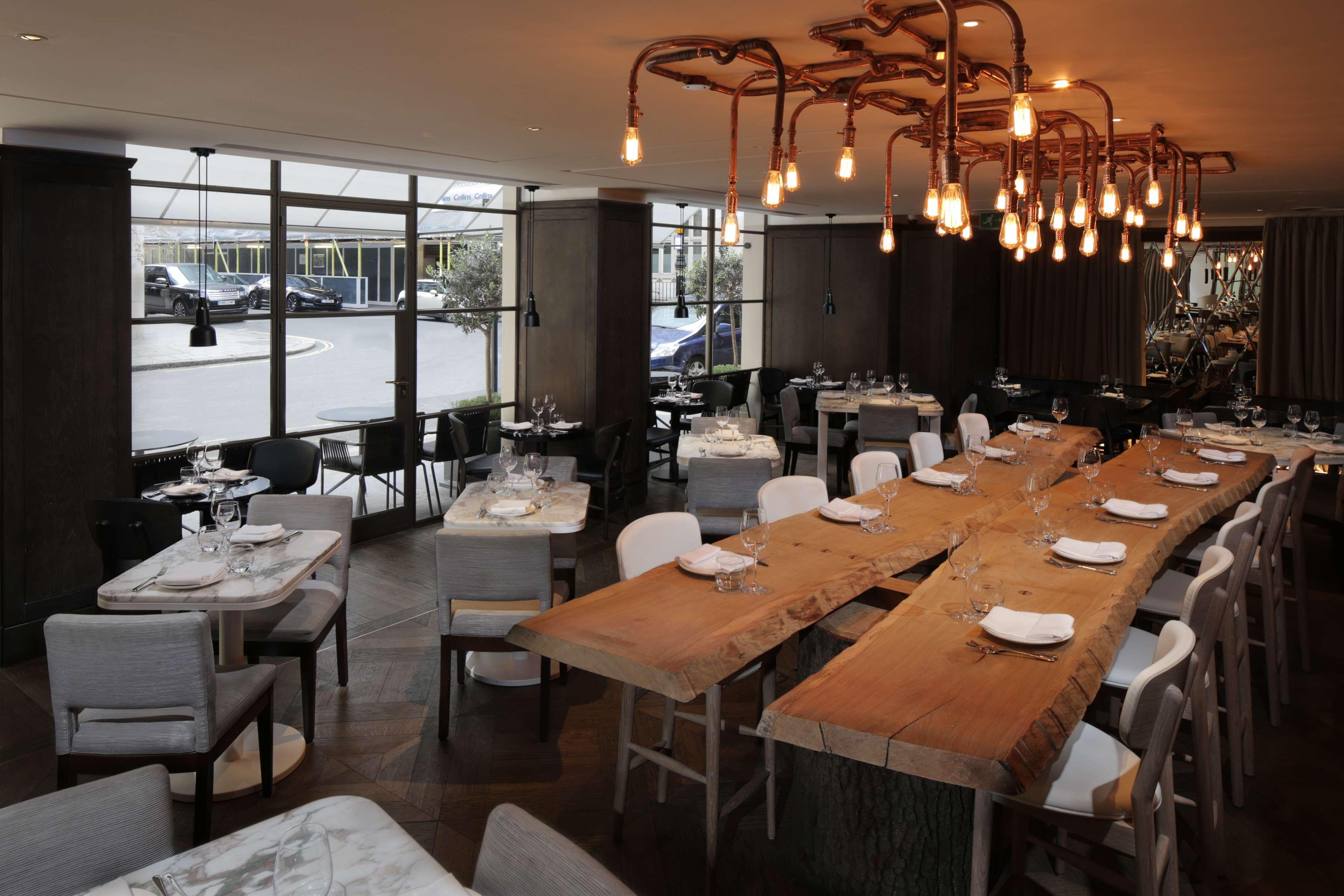 The May Fair, A Radisson Collection Hotel, Mayfair Londra Esterno foto The photo displays a stylish restaurant interior featuring a mix of wooden and upholstered furniture. There's a large wooden table at the center, surrounded by various seating options, including chairs and smaller tables. The room is well-lit, with u