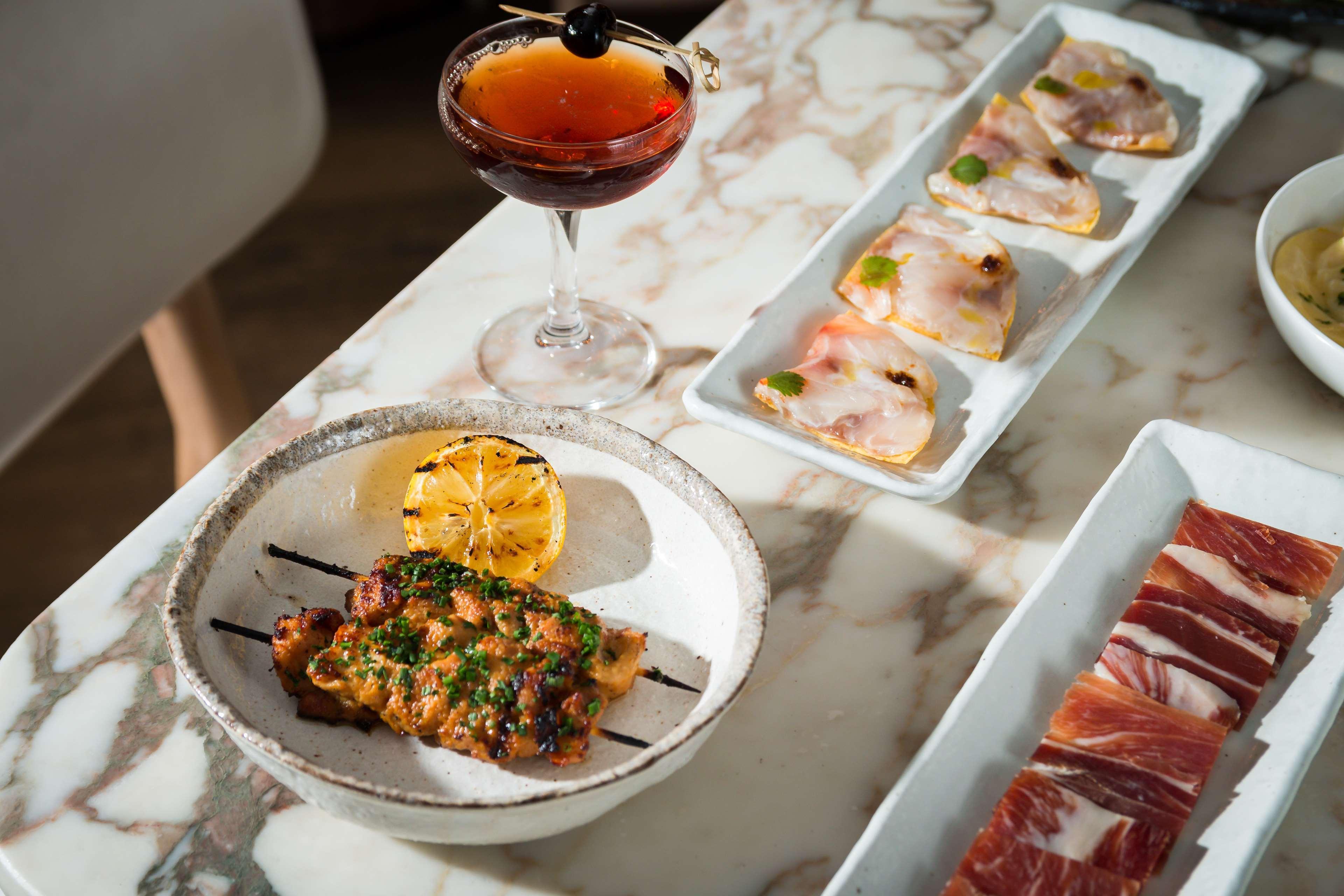 The May Fair, A Radisson Collection Hotel, Mayfair Londra Esterno foto The photo features a beautifully arranged dining table with a variety of food and a drink. In the foreground, there is a dish containing grilled or cooked meat, garnished with herbs and served with a roasted lemon slice. Next to it, a cocktail drink 
