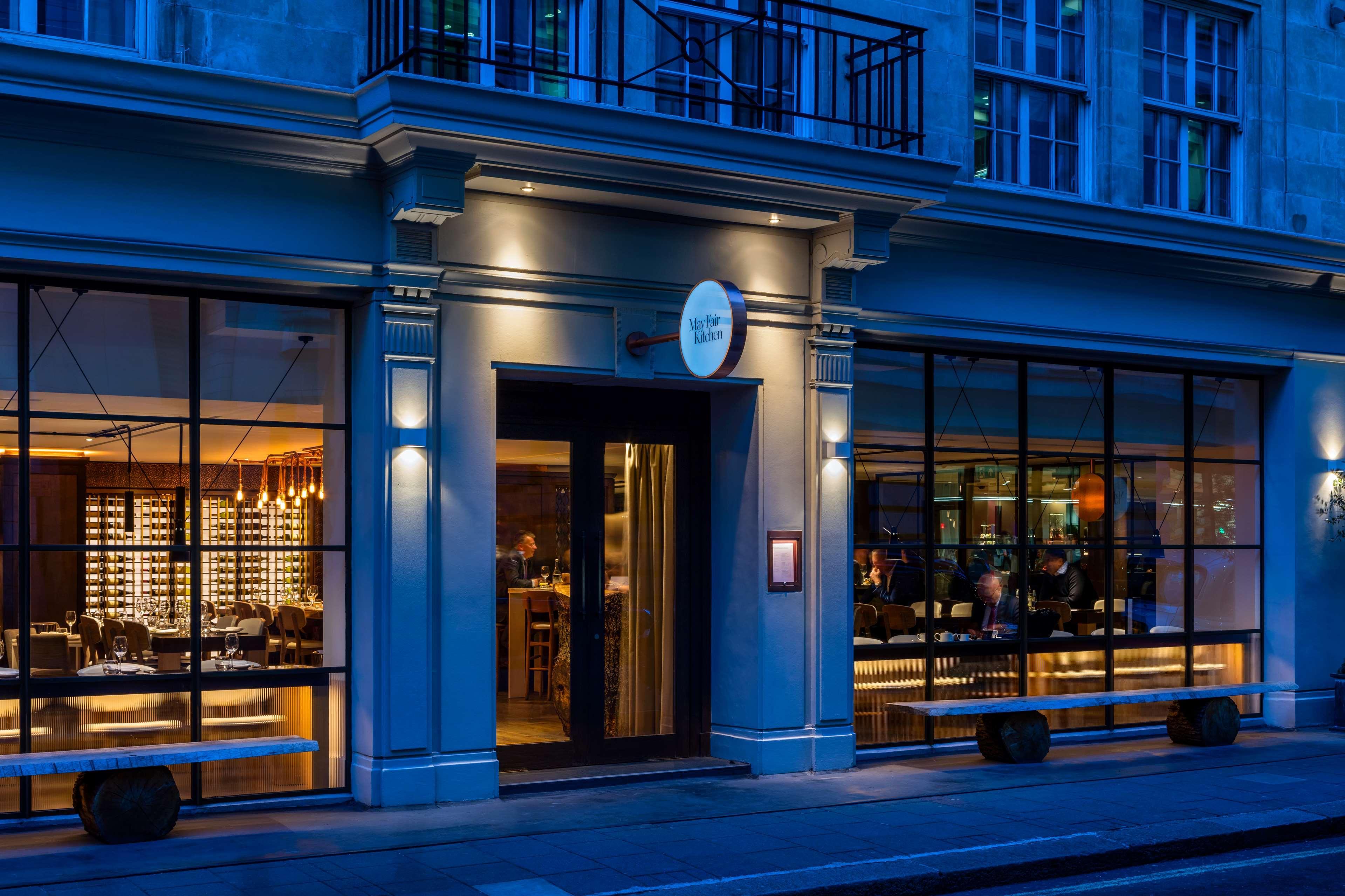 The May Fair, A Radisson Collection Hotel, Mayfair Londra Esterno foto The photo shows the exterior of a stylish restaurant. The entrance features a glass door and large windows, allowing a view inside. The restaurant is illuminated with soft lighting, creating a warm and inviting atmosphere against the evening backdrop