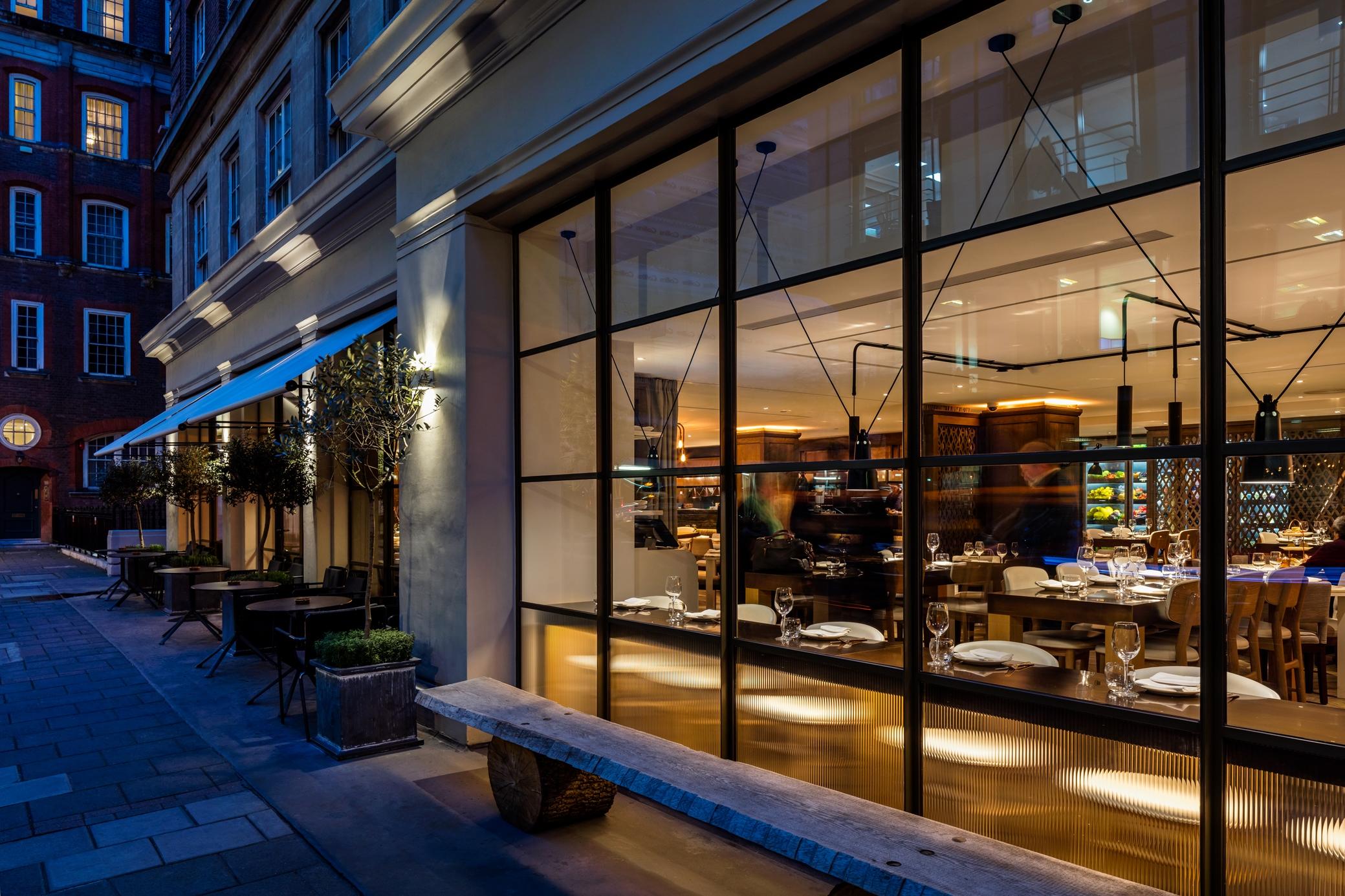 The May Fair, A Radisson Collection Hotel, Mayfair Londra Esterno foto The photo shows the exterior of a restaurant viewed from the outside at night. It features large glass windows that allow a view of the interior, which is warmly lit. Inside, there are tables set for dining, indicating a welcoming atmosphere. Outside