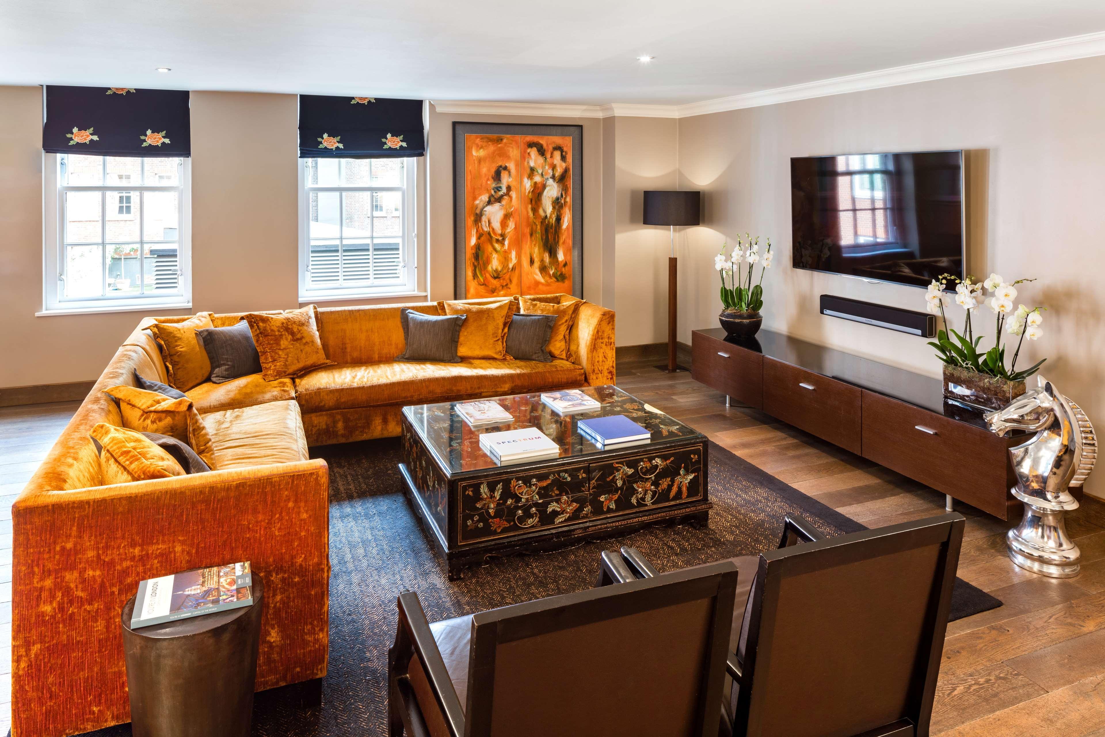 The May Fair, A Radisson Collection Hotel, Mayfair Londra Esterno foto The photo shows a modern living room with a stylish design. It features a large, orange sectional sofa with colorful cushions, positioned around a coffee table that has an ornate design. The floor is wooden, and there's a dark area rug beneath the se