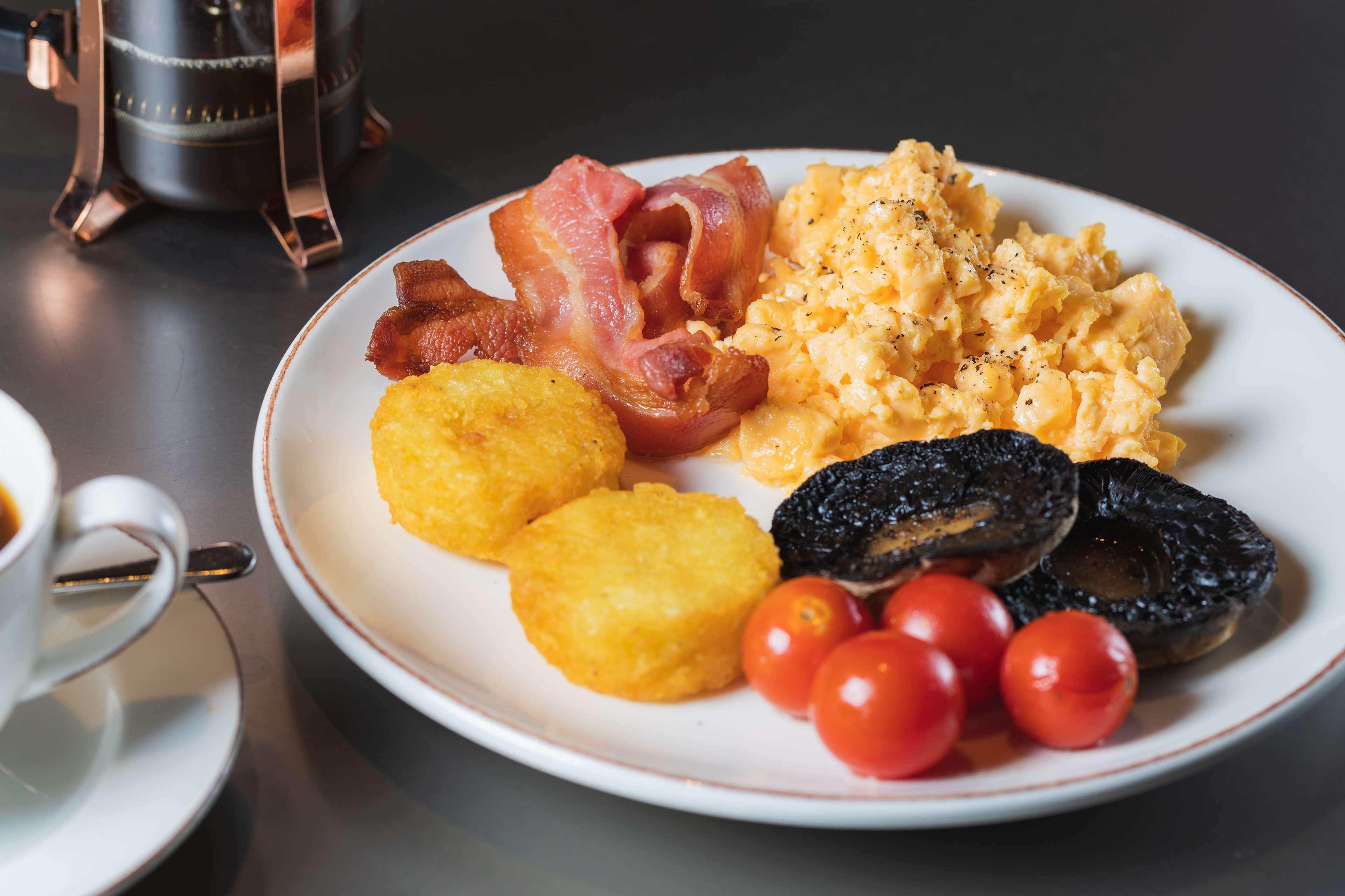 The May Fair, A Radisson Collection Hotel, Mayfair Londra Esterno foto The photo shows a plate of breakfast food that includes scrambled eggs, crispy bacon strips, two golden hash browns, grilled tomatoes, and slices of black pudding. The dish appears to be served alongside a small cup, presumably for coffee or tea. The