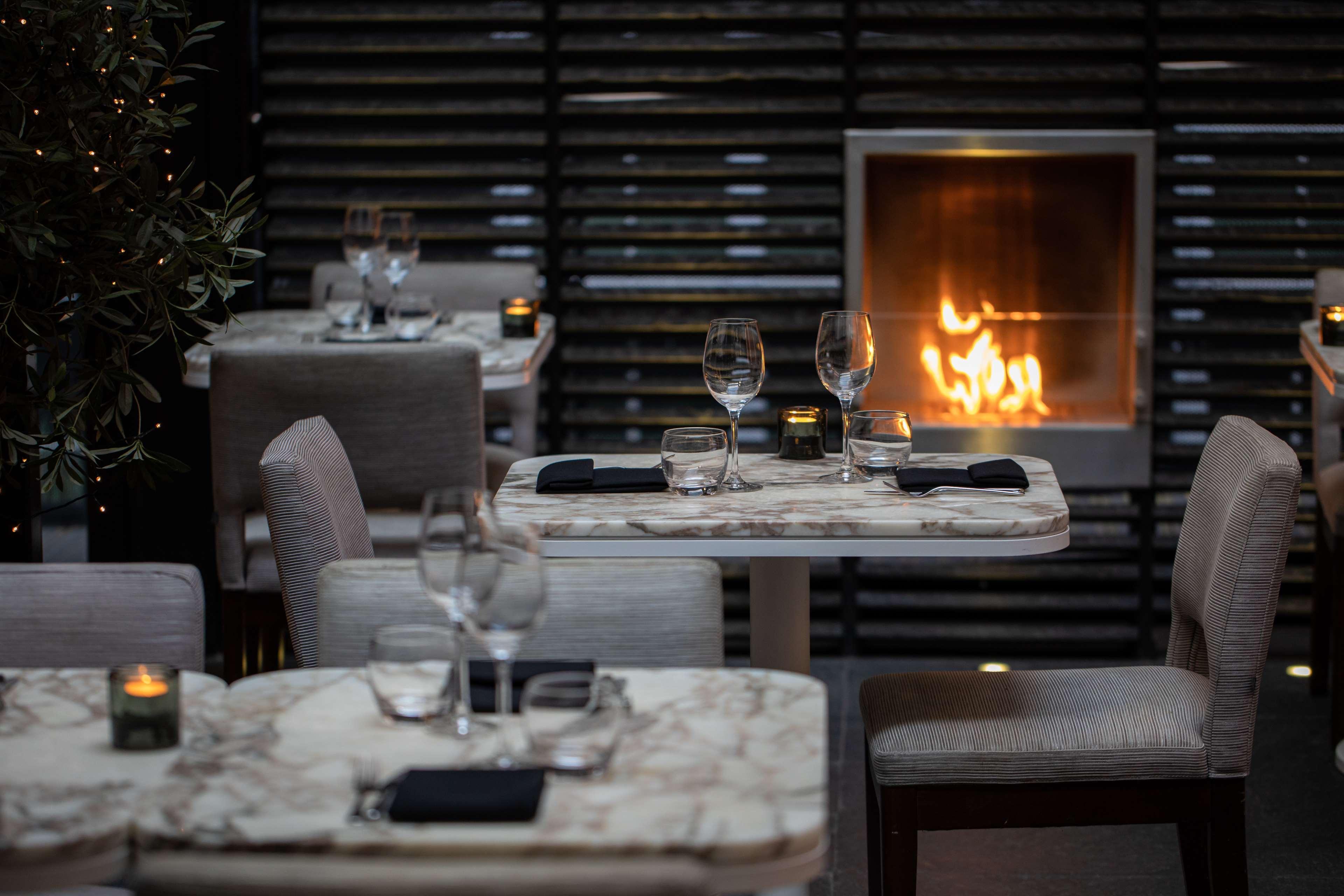 The May Fair, A Radisson Collection Hotel, Mayfair Londra Esterno foto The photo depicts a cozy restaurant interior with elegantly set tables. Each table features a marble top and is equipped with glassware and napkins. There are several tables neatly arranged in the space. In the background, there is a modern fireplace