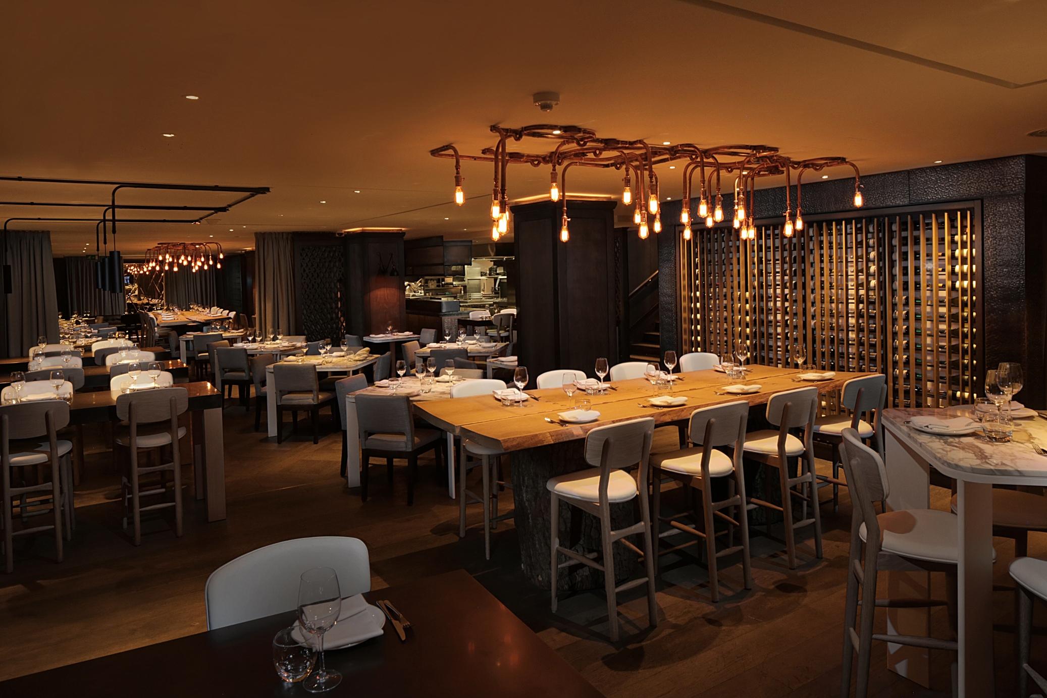 The May Fair, A Radisson Collection Hotel, Mayfair Londra Esterno foto The photo depicts a modern restaurant interior. The space features a combination of wooden and white furniture, with tables set for dining. There is a long, communal-style table at the center, surrounded by high chairs. The lighting is warm, coming f