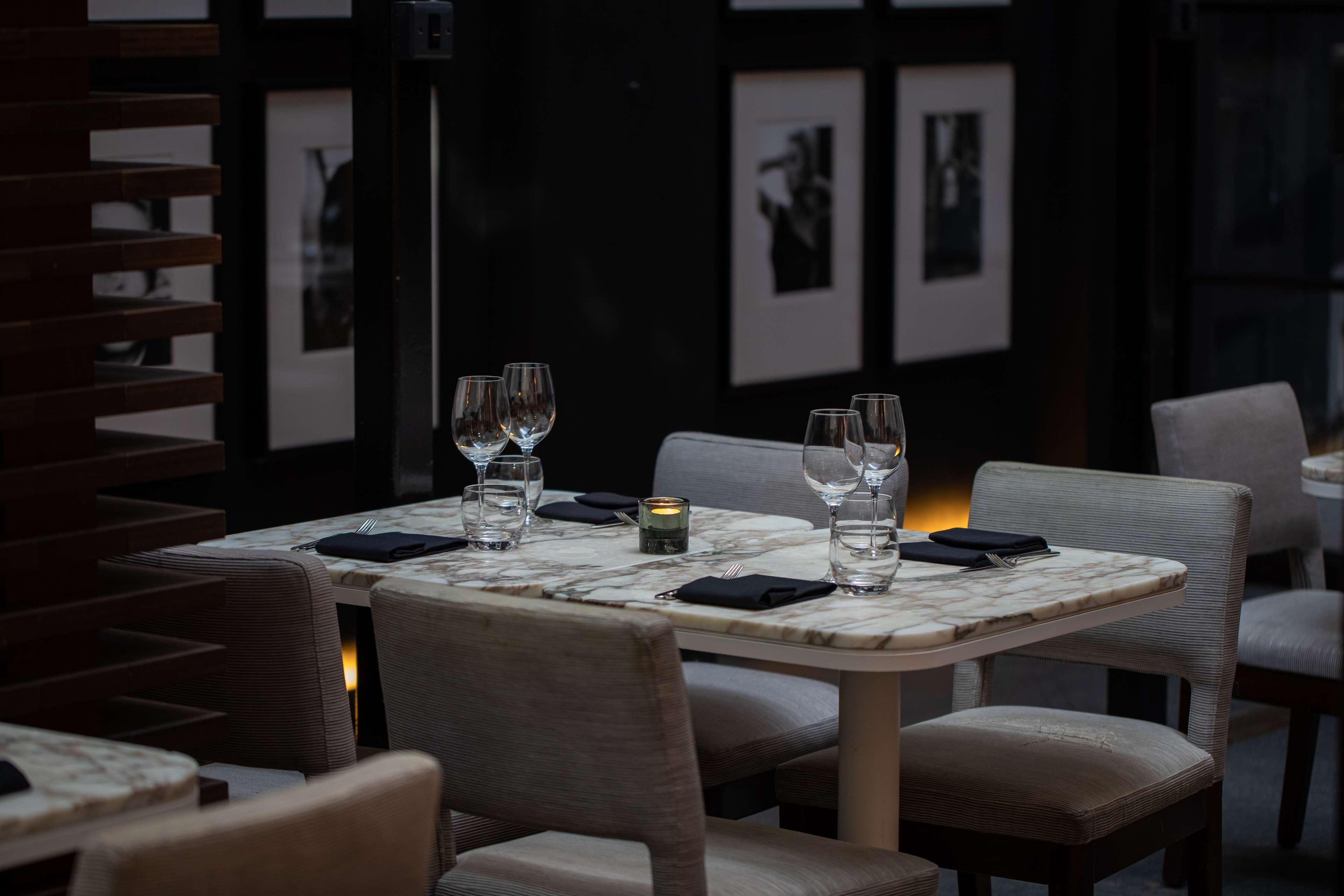 The May Fair, A Radisson Collection Hotel, Mayfair Londra Esterno foto The photo shows a dining area with a stylish and elegant atmosphere. There are two tables set for dining, featuring marble tops and light-colored chairs. Each table has wine glasses and a small candle holder, creating a warm ambiance. In the backgrou