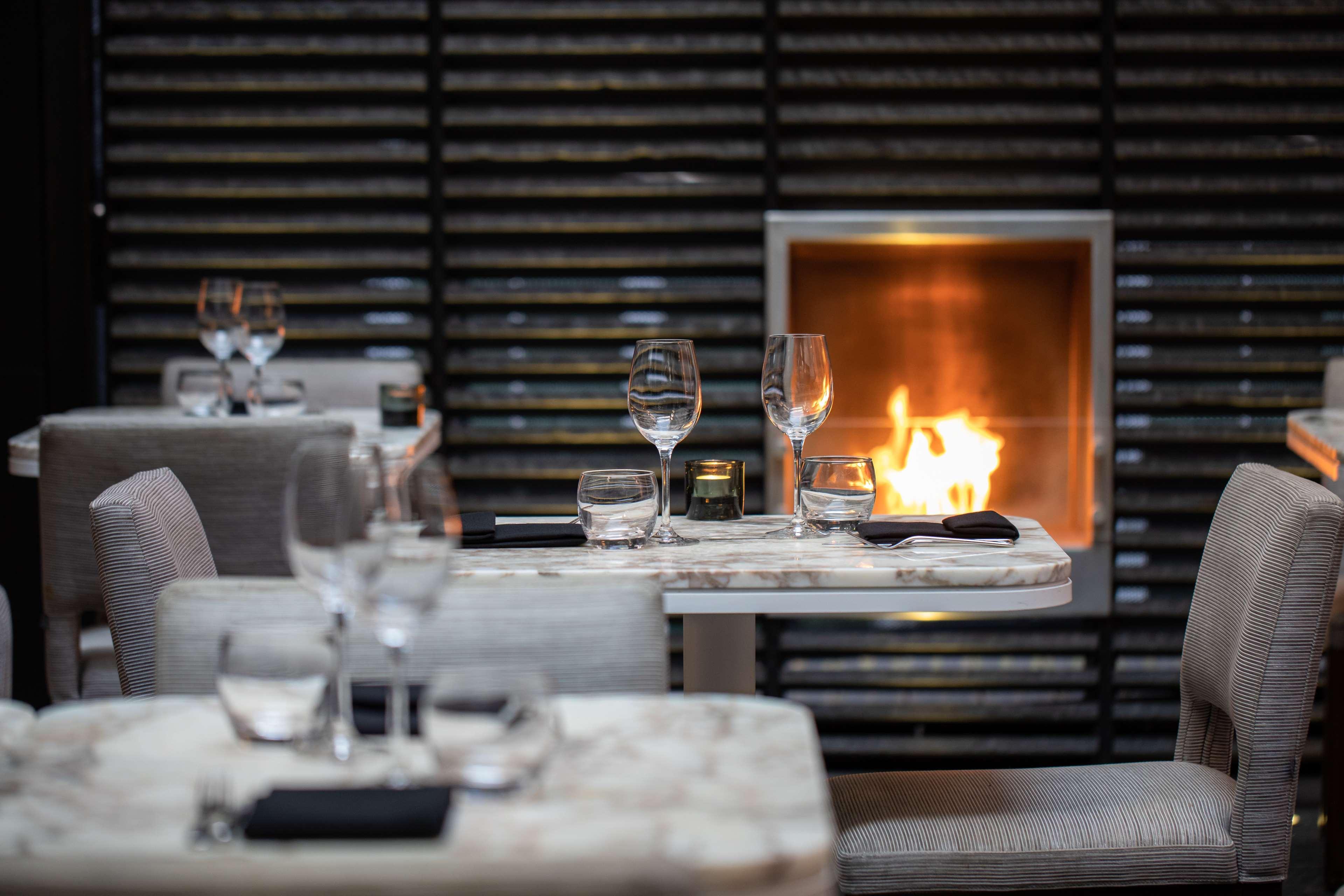 The May Fair, A Radisson Collection Hotel, Mayfair Londra Esterno foto The photo depicts an elegant dining space featuring tables with marble tops, each set with wine glasses. The chairs are upholstered in a soft, neutral fabric. In the background, there is a modern fireplace that adds warmth to the atmosphere with visi
