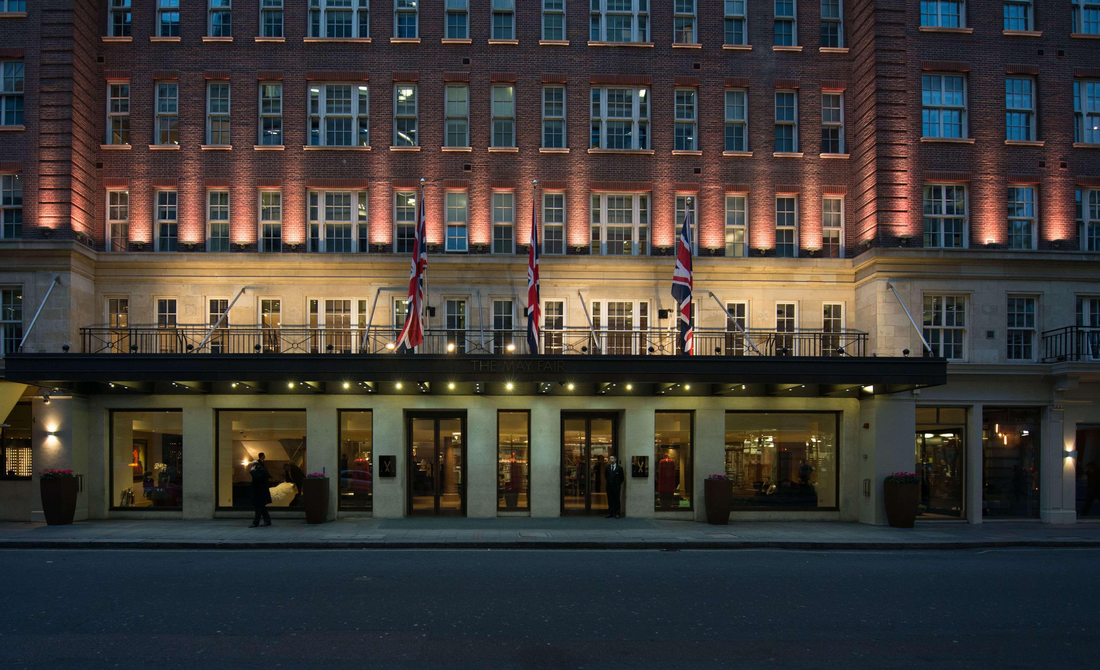The May Fair, A Radisson Collection Hotel, Mayfair Londra Esterno foto The photo depicts a grand hotel exterior at dusk. The building has a classic architectural style, characterized by multiple stories with large windows. The facade is illuminated, highlighting the architectural features. British flags are prominently 
