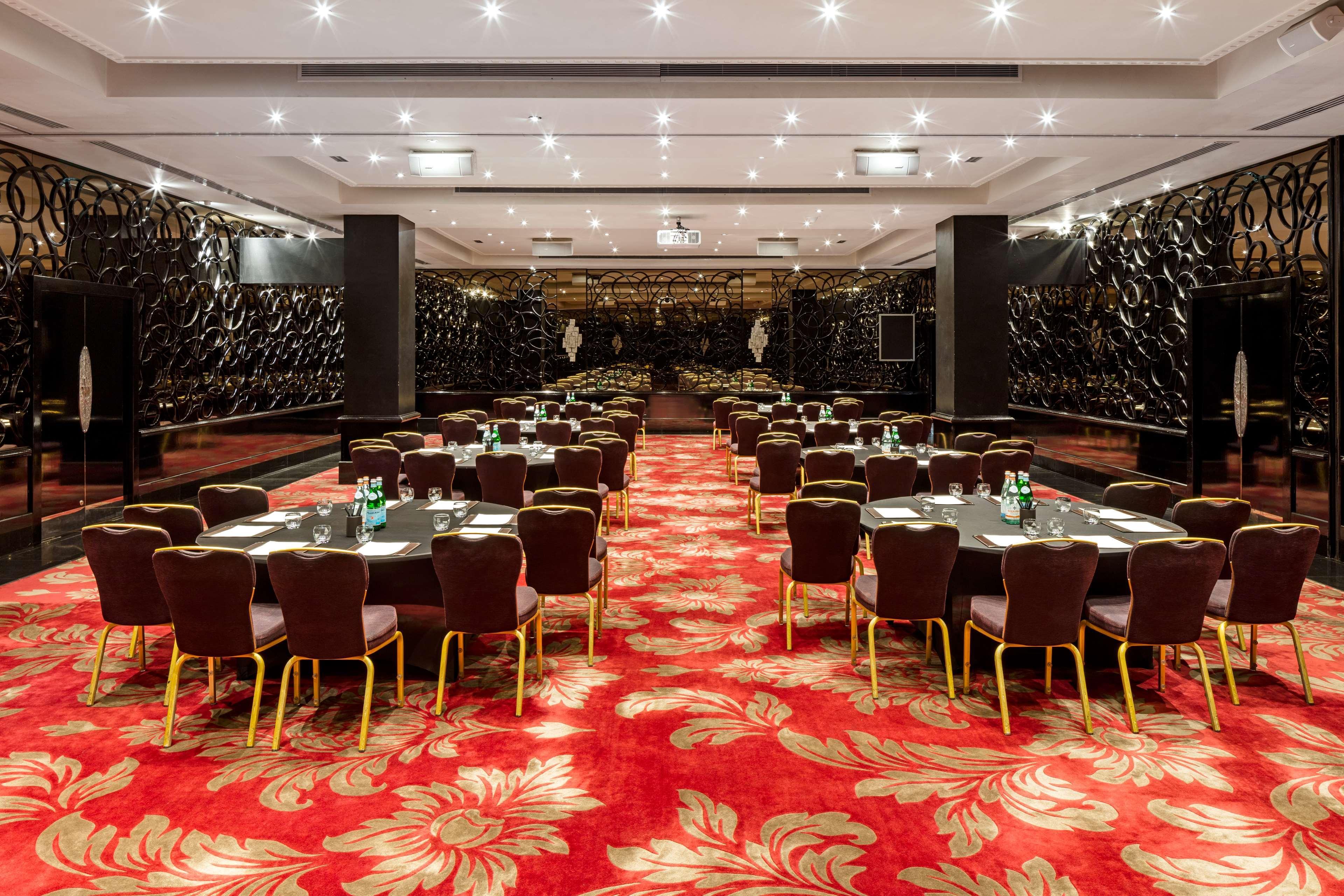 The May Fair, A Radisson Collection Hotel, Mayfair Londra Esterno foto The photo shows a spacious dining or banquet room. It features several round tables arranged neatly with dark chairs. The floor is covered with a vibrant red carpet that has floral patterns. The walls are decorated with a sleek, dark design, adding a