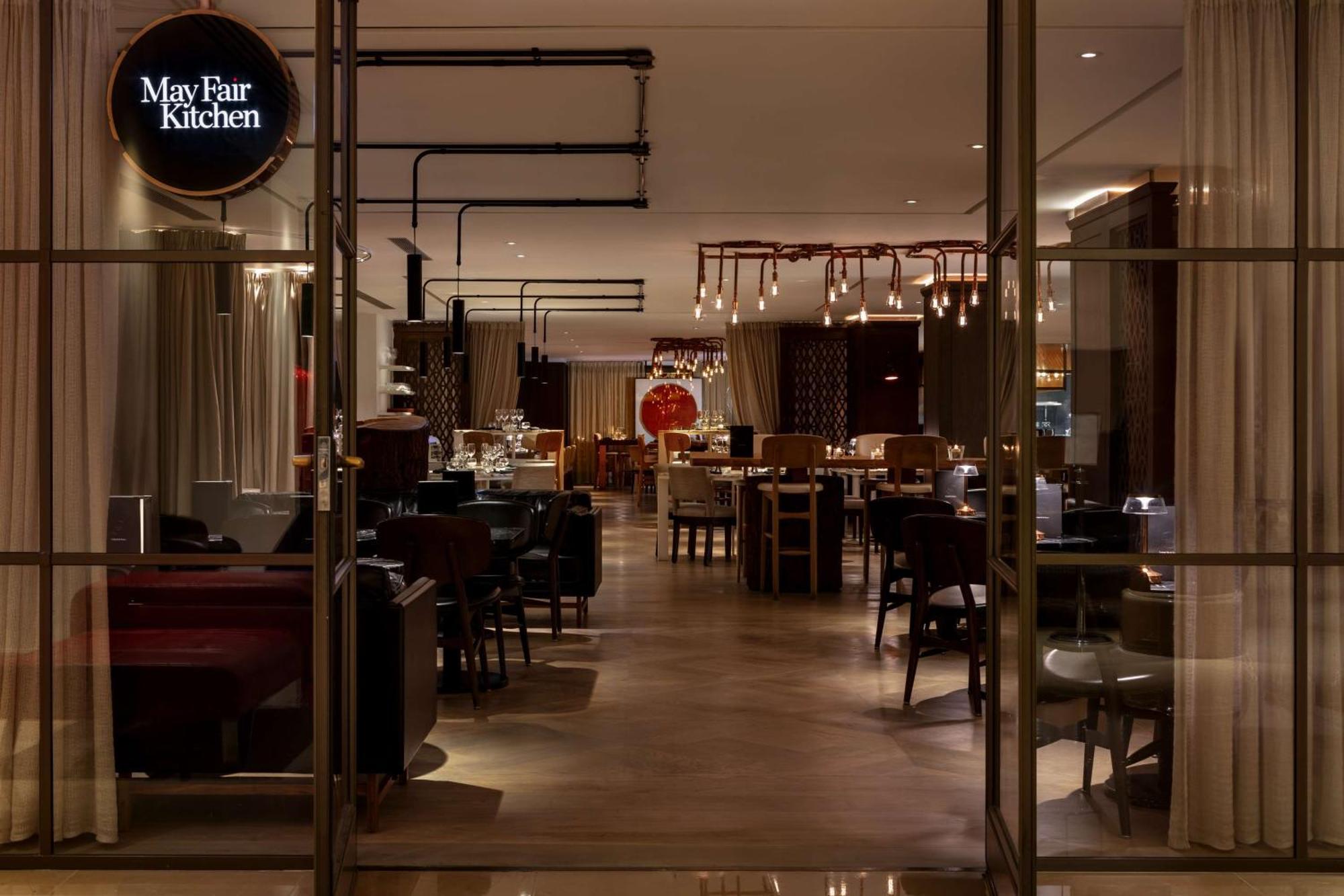 The May Fair, A Radisson Collection Hotel, Mayfair Londra Esterno foto The photo shows the interior of a restaurant called "May Fair Kitchen." The setting features a modern and elegant design, with various tables and chairs arranged throughout the space. There are warm lighting fixtures hanging from the ceiling, and the