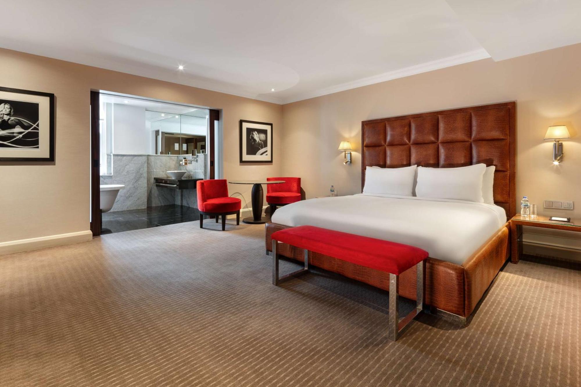 The May Fair, A Radisson Collection Hotel, Mayfair Londra Esterno foto The photo shows a modern hotel room featuring a large, comfortable king-size bed with a beige and brown color scheme. There are two red accent chairs positioned near the bed, adding a pop of color. A small red bench is placed at the foot of the bed. 