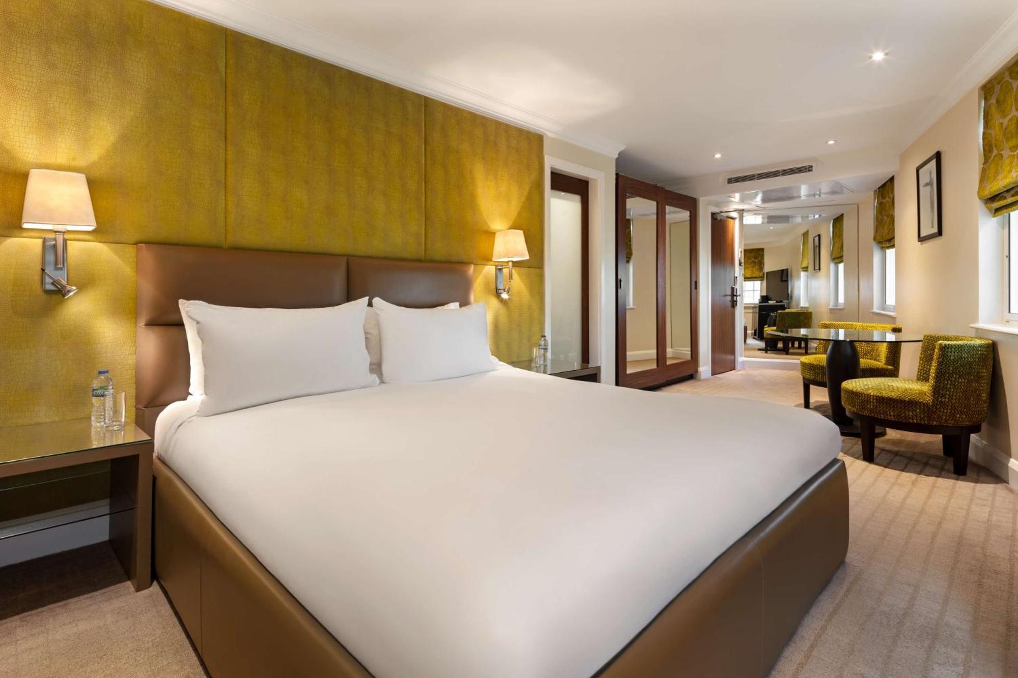 The May Fair, A Radisson Collection Hotel, Mayfair Londra Esterno foto The photo shows a modern hotel room featuring a large bed with a neatly made white duvet and pillows. The walls are accented with a soft yellow color, enhancing the inviting atmosphere. On either side of the bed, there are bedside tables with lamps. 