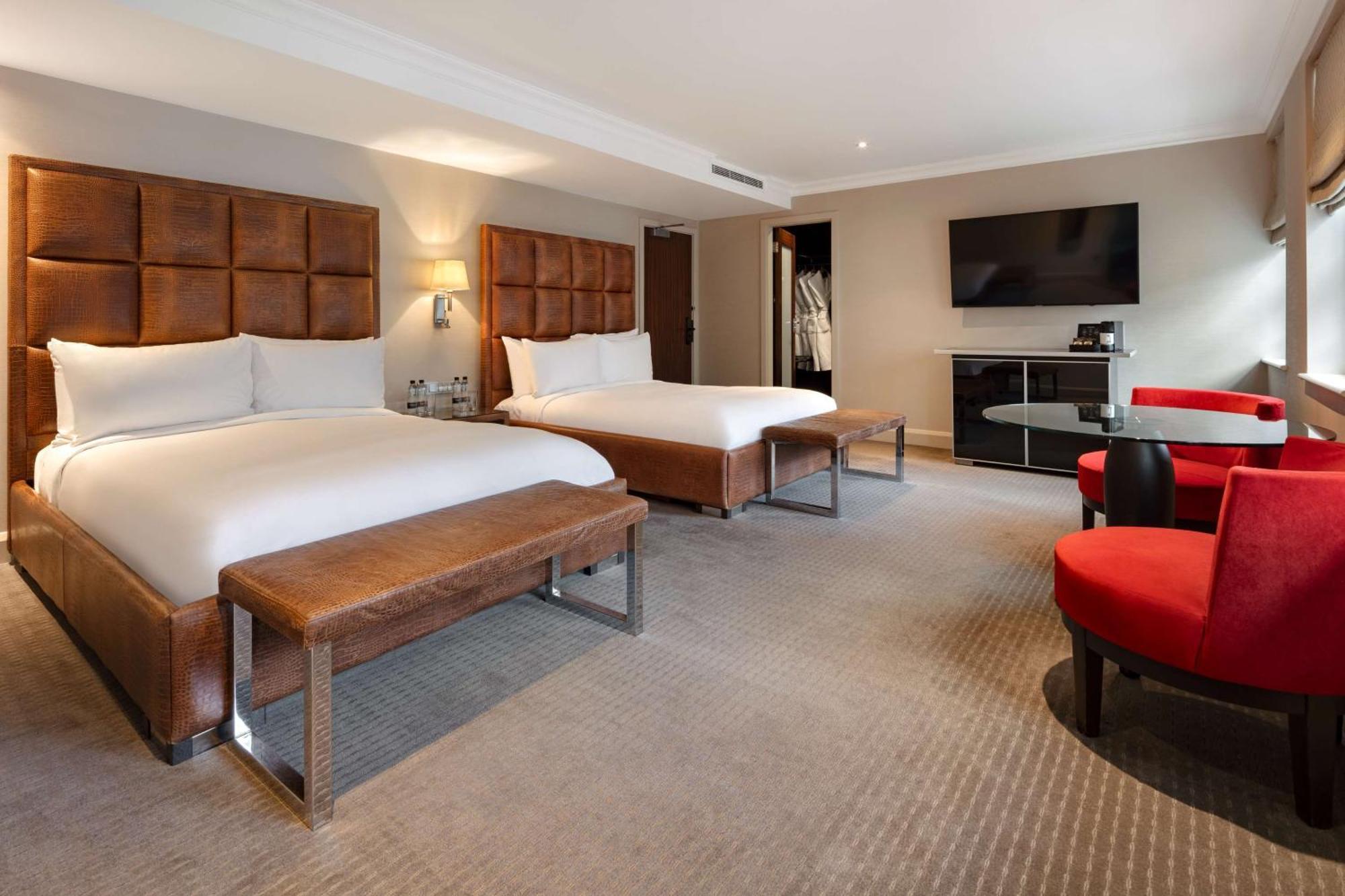 The May Fair, A Radisson Collection Hotel, Mayfair Londra Esterno foto The photo shows a modern hotel room with two beds. Each bed is made with white linens and has a large, upholstered headboard. There is a bench at the foot of each bed. To the right, there are two red chairs around a small glass table. A flat-screen T