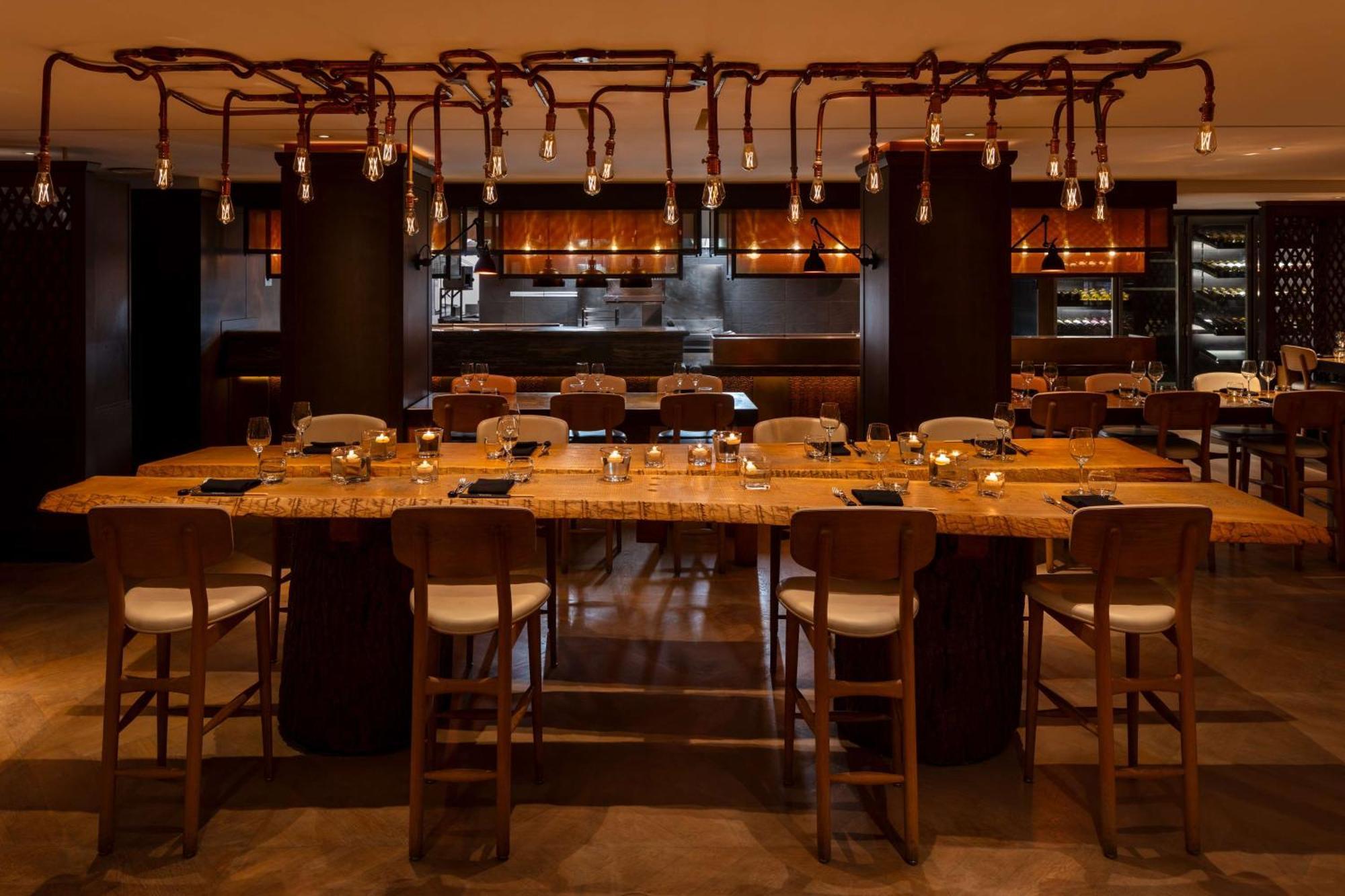 The May Fair, A Radisson Collection Hotel, Mayfair Londra Esterno foto The photo shows a modern restaurant interior featuring a long wooden dining table with several high chairs around it. The table is set with plates and glasses, suggesting it is ready for guests. Above the table, there are unique light fixtures that r