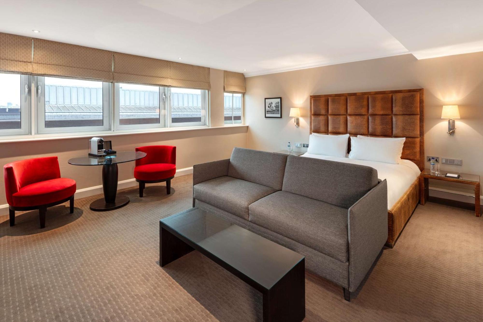 The May Fair, A Radisson Collection Hotel, Mayfair Londra Esterno foto The photo shows a modern hotel room featuring a comfortable setup. There is a large double bed with a headboard, some decorative artwork on the wall, and two windows allowing natural light to enter. In the room, there's a gray sofa facing a dark coff