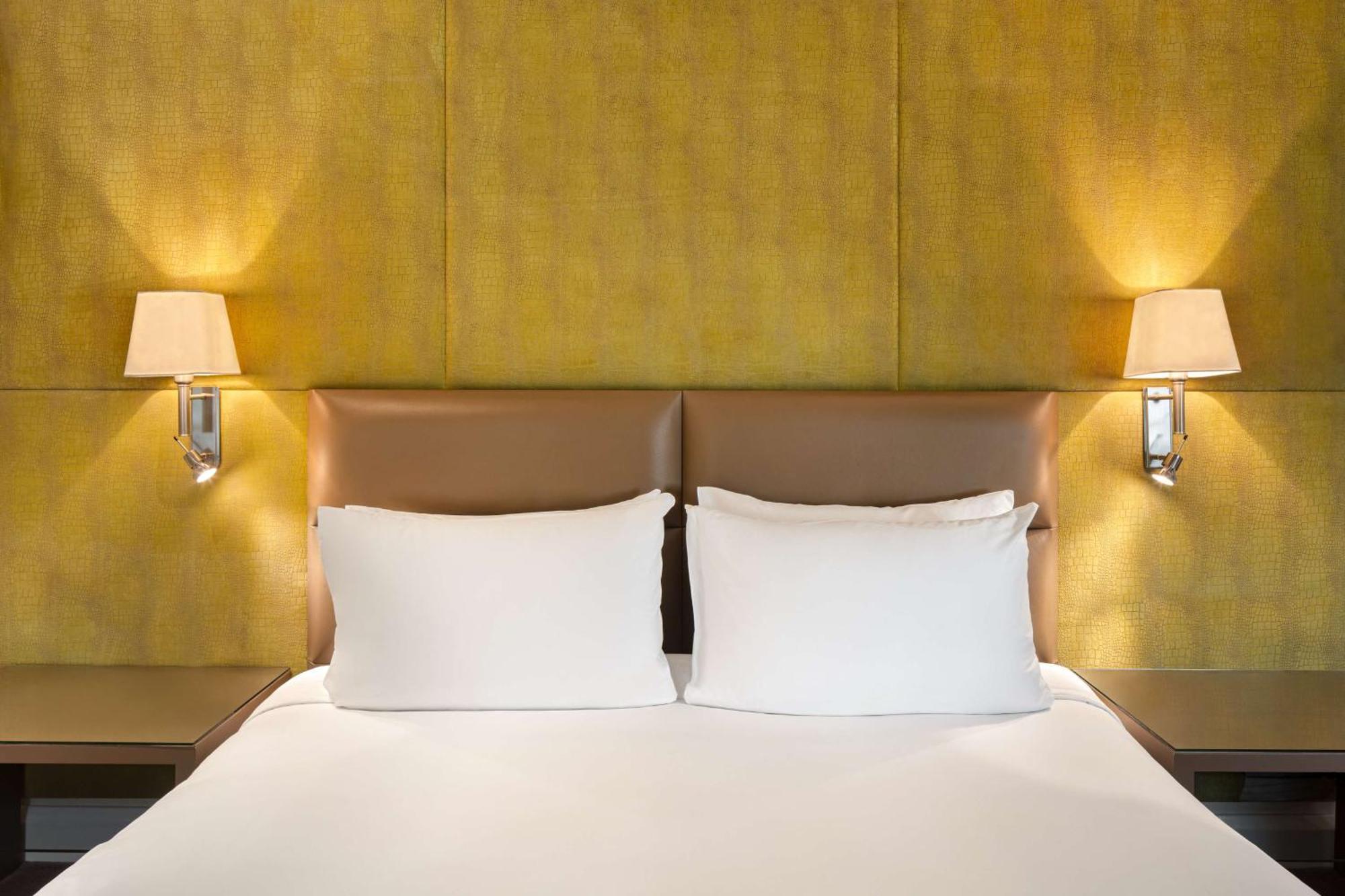 The May Fair, A Radisson Collection Hotel, Mayfair Londra Esterno foto The photo shows a cozy bedroom setting featuring a large bed with two white pillows. The headboard has a smooth, light brown leather material, and there are two wall-mounted lamps on either side of the bed, illuminating a textured yellow wall behind 