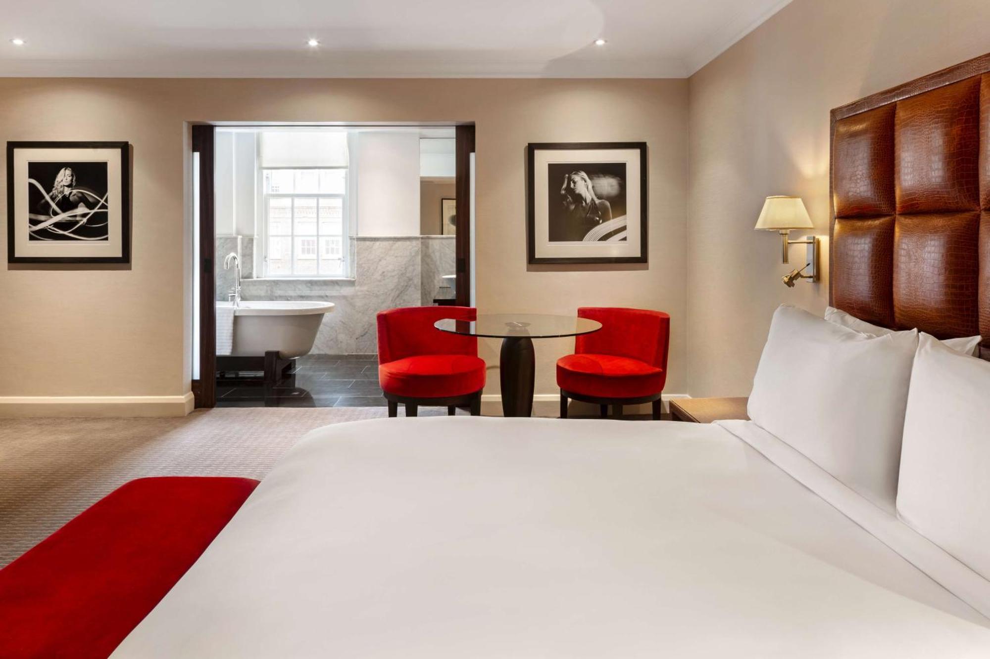 The May Fair, A Radisson Collection Hotel, Mayfair Londra Esterno foto The photo shows a hotel room featuring a large bed with white linens and a red accent throw. In the background, there are two modern red chairs around a small glass table. The wall displays framed black and white photographs. To the side, there is an