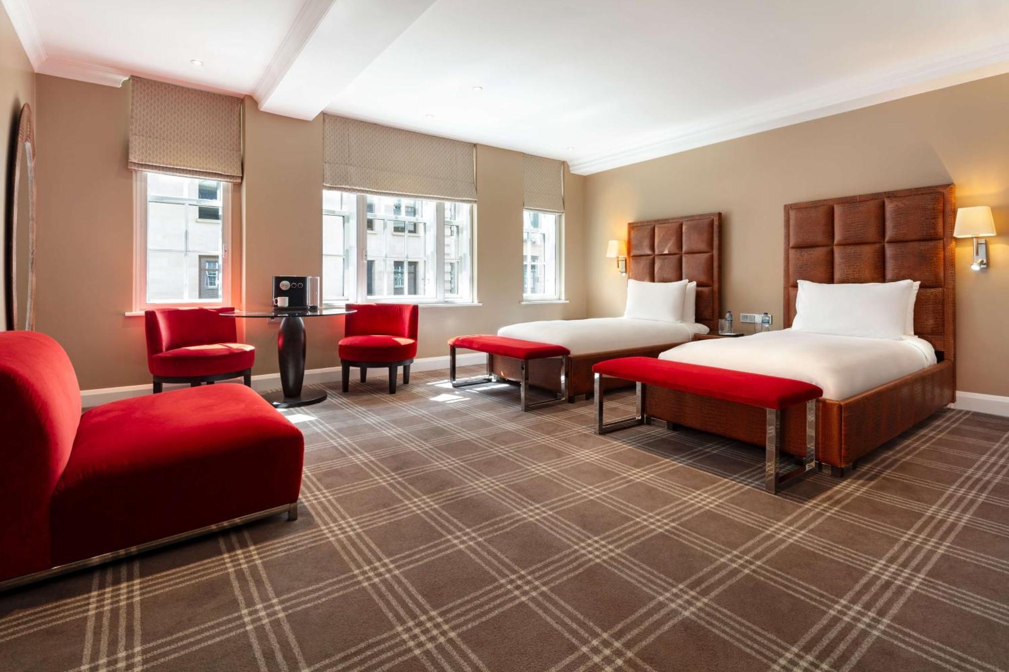 The May Fair, A Radisson Collection Hotel, Mayfair Londra Esterno foto The photo shows a modern hotel room featuring two beds with white bedding and brown headboards. There are two red benches at the foot of each bed. A red upholstered chair and a small round table are positioned near the window, which allows natural li