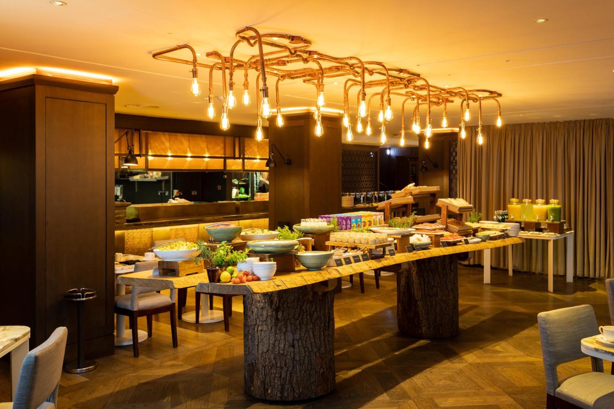The May Fair, A Radisson Collection Hotel, Mayfair Londra Esterno foto The photo shows a stylish dining area with a buffet setup. In the center, there is a long wooden table made from a tree trunk, displaying various dishes and food items. The setting features an elegant lighting fixture made of exposed bulbs that resem