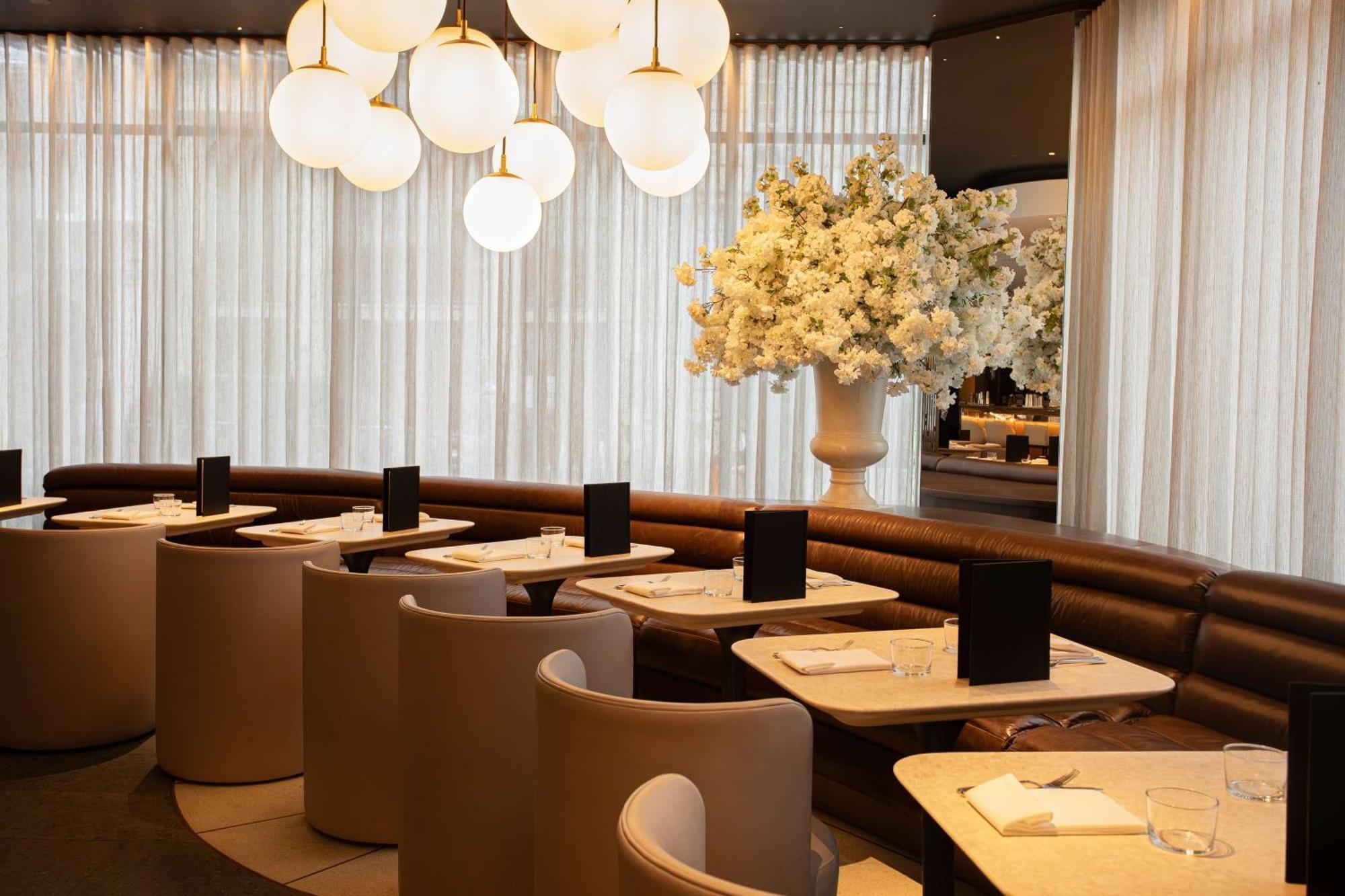 The May Fair, A Radisson Collection Hotel, Mayfair Londra Esterno foto The photo shows a modern restaurant interior. It features stylish seating arrangements with curved booths and elegant tables. The lighting is soft, provided by large, round pendant lights hanging from the ceiling. A large floral arrangement in a whit