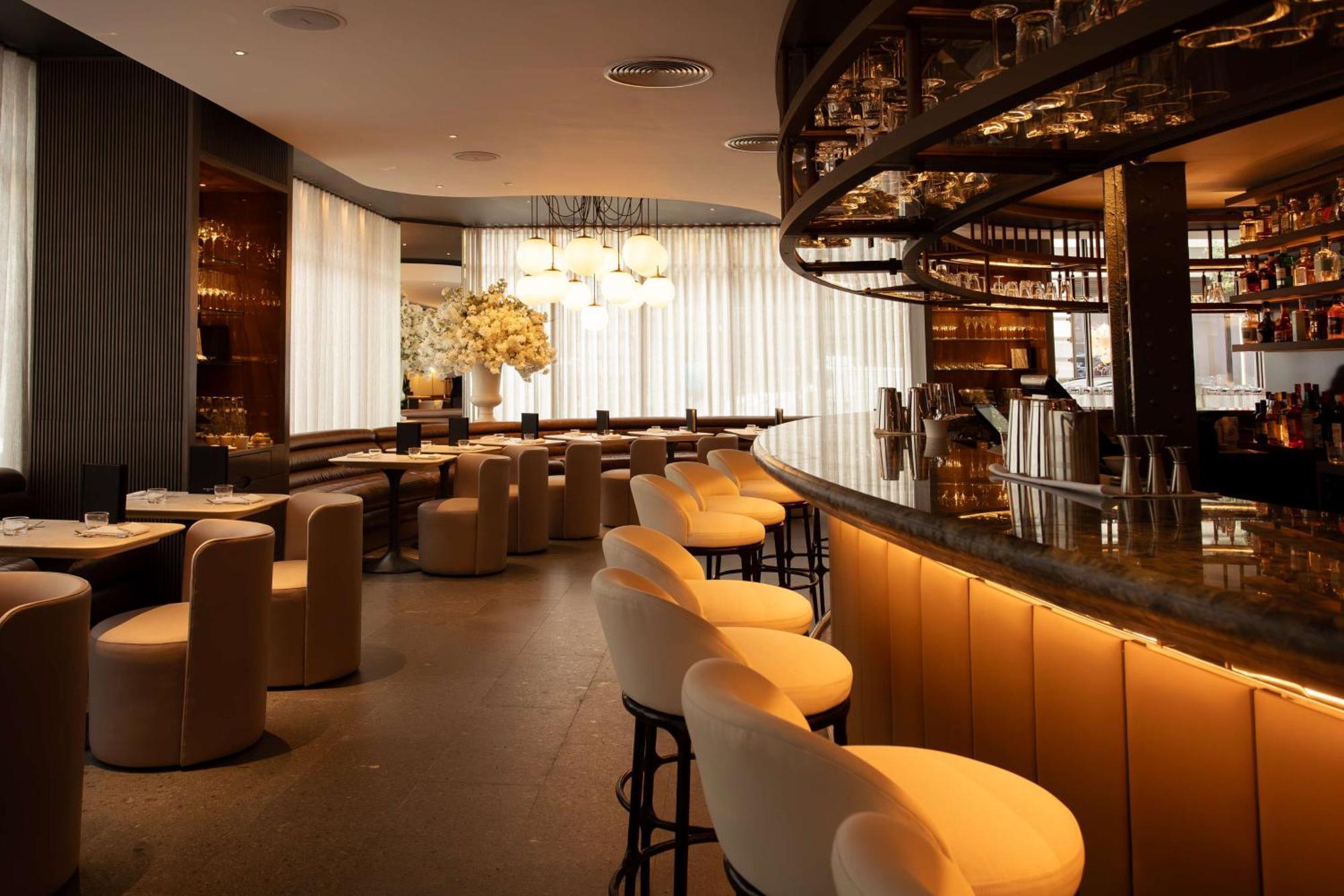 The May Fair, A Radisson Collection Hotel, Mayfair Londra Esterno foto The photo shows the interior of a sleek and modern bar or restaurant. There is a curved bar counter with several stylish high stools in front of it. The seating area includes plush, rounded chairs arranged around circular tables. The design features 